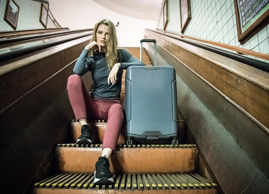Jonge vrouw zit op trap met blauwe koffer, in smalle hal met houten wanden en glazen ramen.